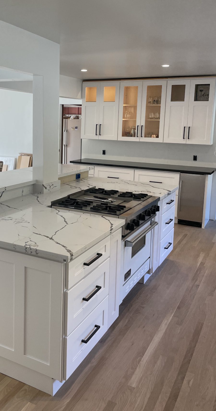 A kitchen near completion: white shaker cabinets with black pulls, glass-front uppers lit from inside, a white quartz island with gray veining around a stainless range, and an oak floor.